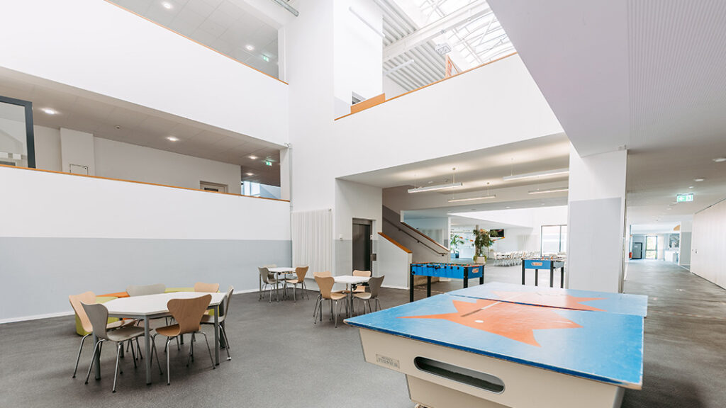 Helle Aufenthaltszone mit mehreren Tischen und Stühlen, Tischtennisplatte mit blauem Belag und orangem Muster, Treppe und Galerie im Hintergrund.