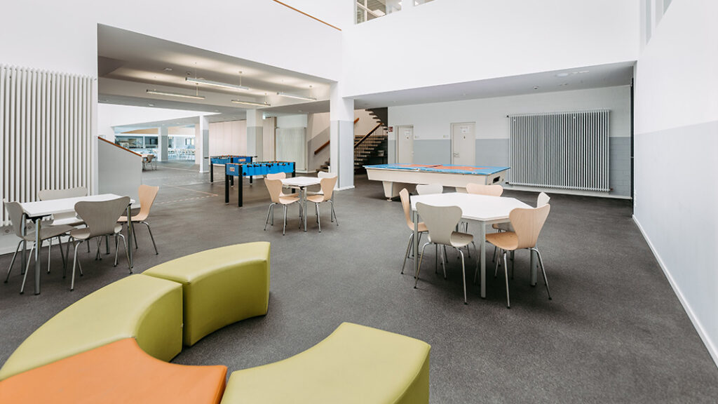 Großer heller Aufenthaltsraum mit mehreren runden Tischen, Stühlen, einem grünen und orangen Sitzkreis sowie einem Kicker und einem Billardtisch.