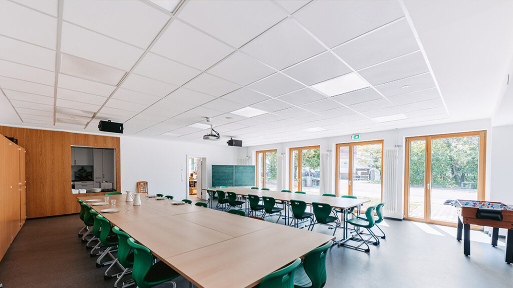 Großer Aufenthaltsraum mit langen Tischen, grünen Stühlen, großer Fensterfront und Kicker-Tisch am rechten Rand.