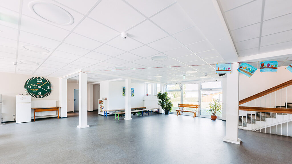 Helle, großzügige Aula einer Grundschule mit großen Fenstern, Sitzbänken, Pflanzen und einer großen grünen Wanduhr.