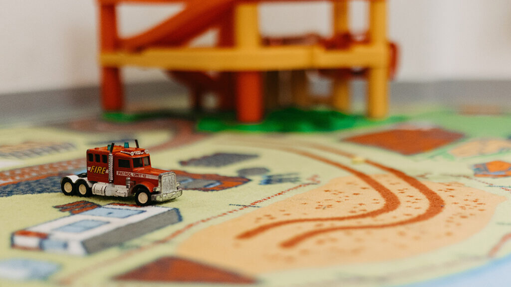 Rotes Spielzeug-Feuerwehrauto auf bunter Spielmatte mit Straßen- und Gebäudemotiven.