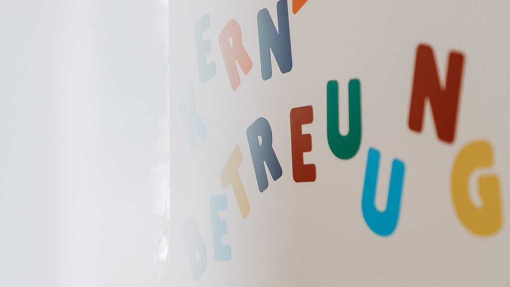 Bunte, einzeln angebrachte Buchstaben an einer weißen Wand, die das Wort 'KERNZEITBETREUUNG' bilden.
