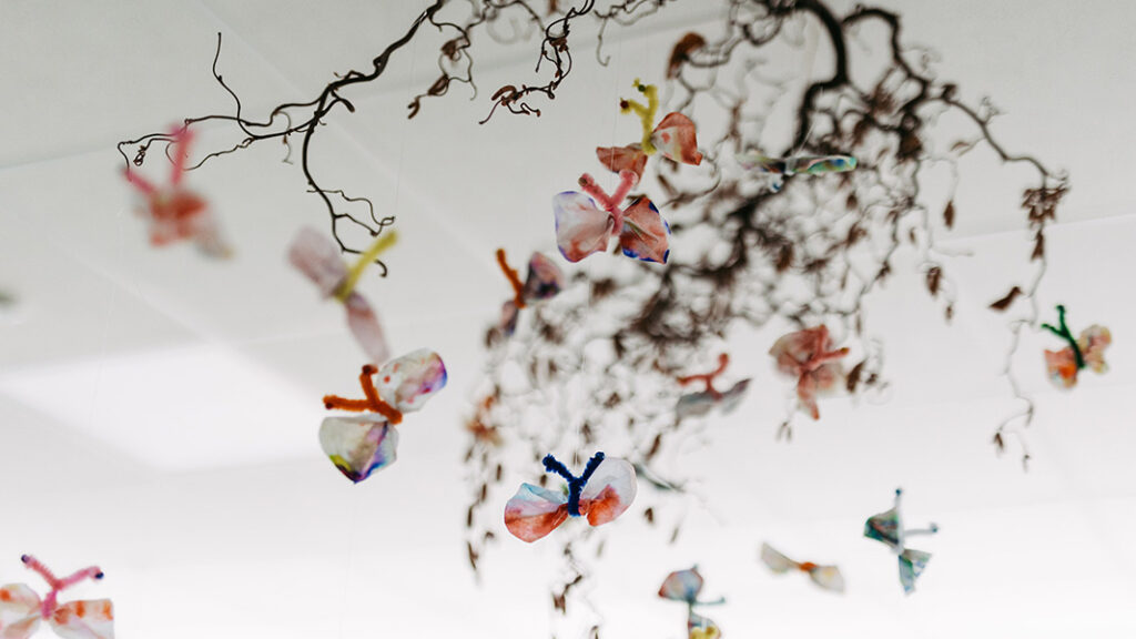 Mehrere bunte Schmetterlinge aus Papier hängen an dünnen, trockenen Zweigen an der Zimmerdecke.