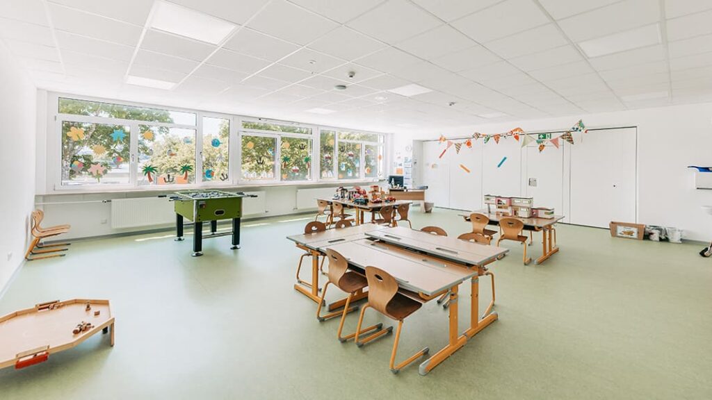 Helles Klassenzimmer mit mehreren Tischen und Stühlen, Kicker, Fenster mit Blick ins Grüne und bunte Girlanden an der Wand.