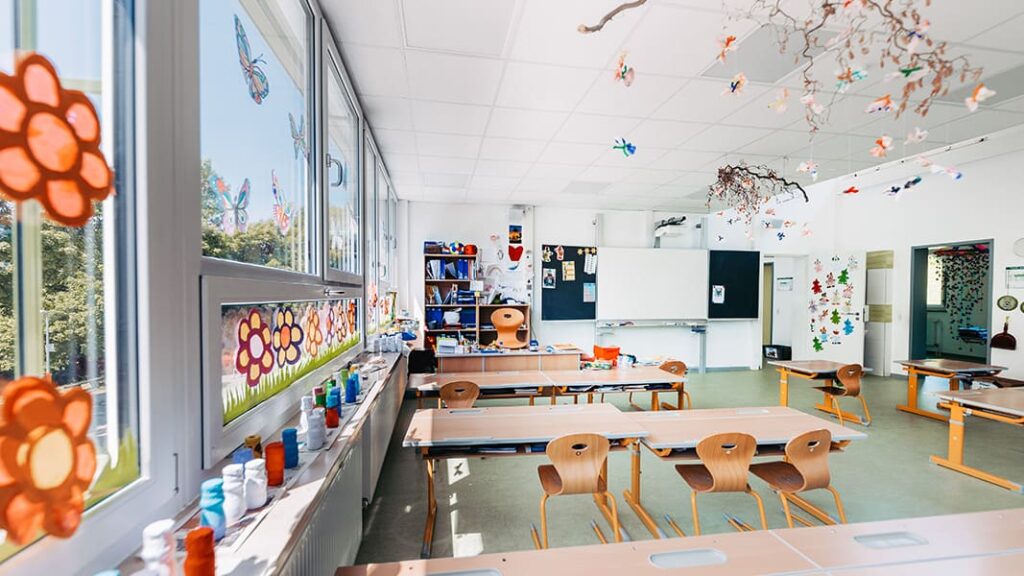 Helles Klassenzimmer mit Holztischen und -stühlen, Fenster mit Blumen- und Schmetterlingsdekoration, bunte Bastelarbeiten an der Decke.