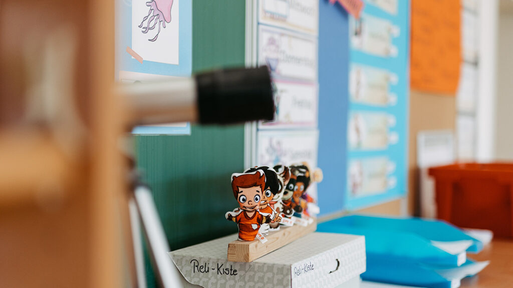 Mehrere kleine Figuren mit lachenden Gesichtern stehen auf einem Holzblock mit der Aufschrift 'Reli-Kiste' vor einer Tafel und bunten Plakaten in einem Klassenzimmer.