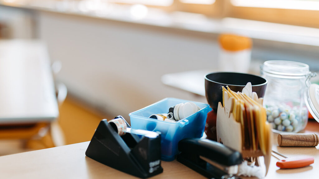Schreibtisch mit schwarzem Klebebandabroller, blauem Behälter mit Büroklammern, schwarzer Tasse, Briefumschlägen und Glas mit bunten Perlen.