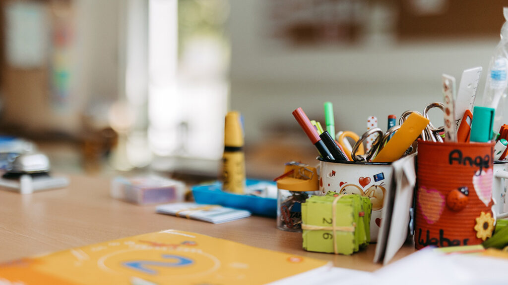 Nahaufnahme von bunten Schreibutensilien in Bechern auf einem Holztisch, darunter Stifte, Marker und Scheren.
