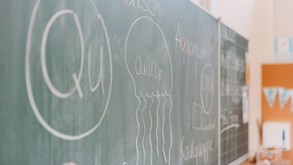 Grüne Schultafel mit Kreidezeichnungen, darunter ein Kreis mit 'Qu' und eine Qualle mit dem Wort 'Qualle' darin.