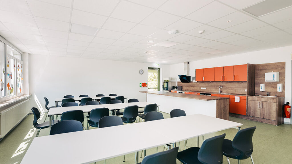 Heller, moderner Klassenraum mit mehreren weißen Tischen und schwarzen Stühlen, mit einer orangefarbene Küche an der Rückwand.