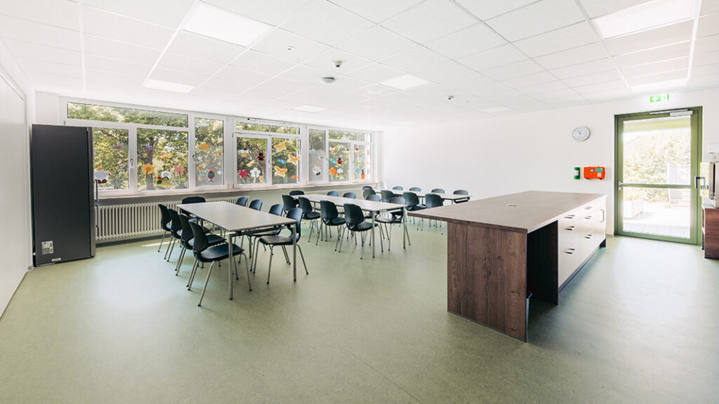 Großer heller Raum mit mehreren Tischen und Stühlen, Fenster mit bunten Aufklebern, Küchentheke mit Schubladen und Tür mit Notausgangsschild.