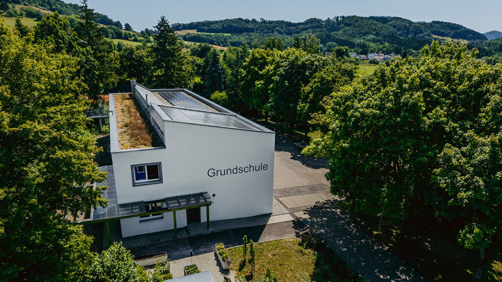 Moderner, weißer Schulgebäudetrakt mit Flachdach und Solarzellen, umgeben von Bäumen und Hügeln, mit dem Schriftzug 'Grundschule'.