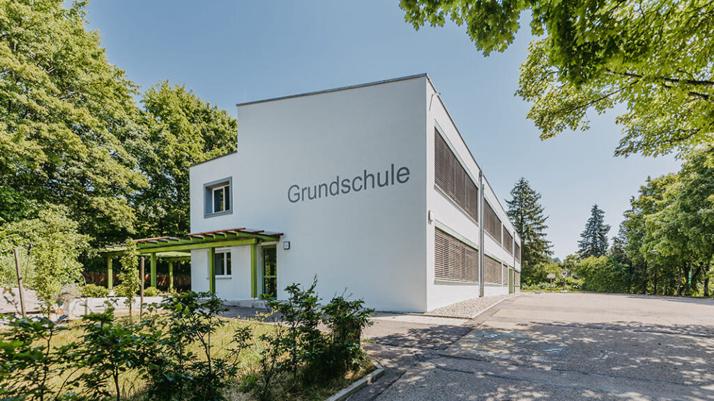 Rechteckiges, weißes Schulgebäude mit der Aufschrift 'Grundschule' an der Fassade, umgeben von Bäumen und Grünflächen.
