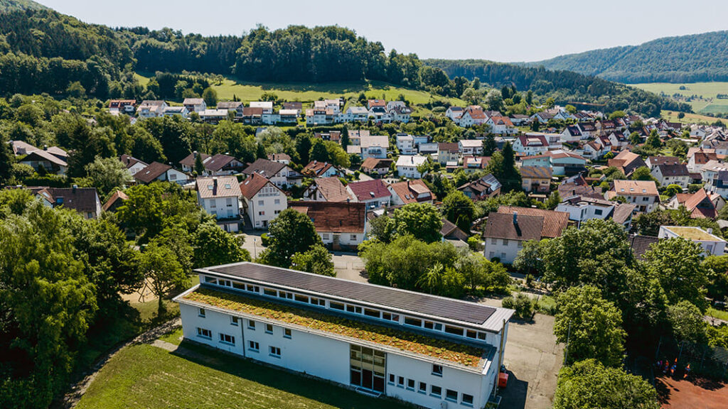 Luftaufnahme einer Grundschule mit Flachdach und Solarpaneelen, umgeben von Bäumen und Wohnhäusern in einer ländlichen Gegend.