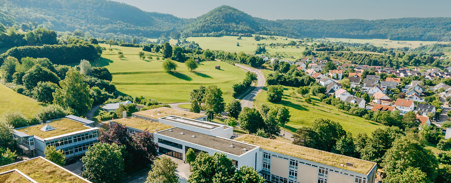 Blick aus der Luft auf eine grüne Landschaft mit grünen Feldern, Bäumen, Wohnhäsuern und einem Teil des Schulkomplexes der Messelbergschule im Vordergrund unter blauem Himmel.