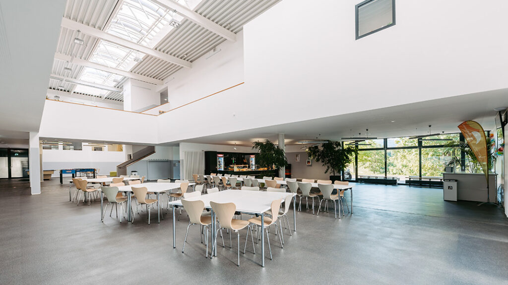 Großer heller Speisesaal mit mehreren rechteckigen Tischen und Stühlen, großen Fenstern und Oberlichtern.