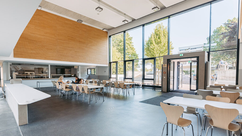 Großer heller Speisesaal mit hohen Fenstern, mehreren Tischen und Stühlen, einige Personen sitzen und essen.