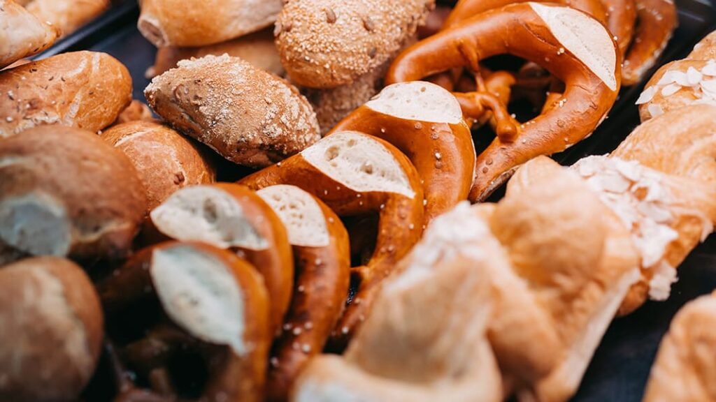Verschiedene Brotsorten, darunter Laugenbrezeln mit Schnitt, Körnerbrötchen und helle Brötchen, nah beieinander liegend.