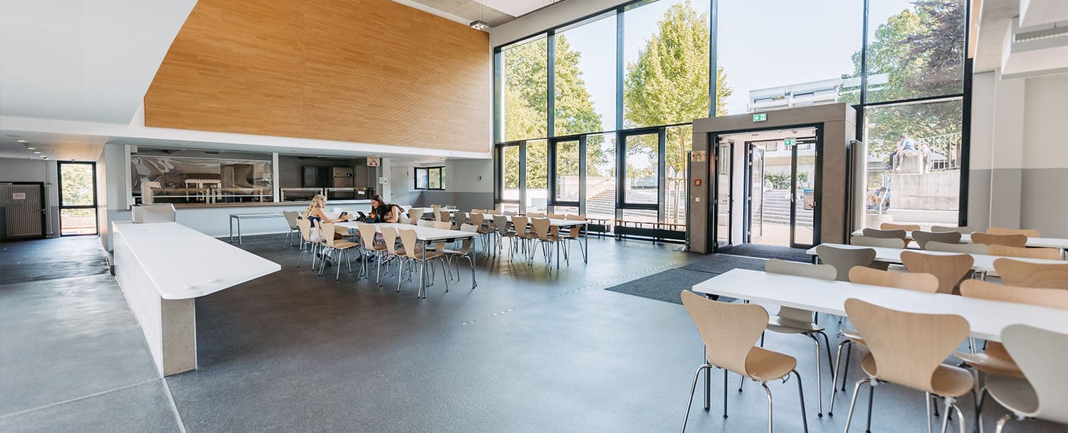 Helle, moderne Mensa mit großen Fenstern, mehreren Tischen und Stühlen, zwei Personen sitzen an einem Tisch und unterhalten sich.