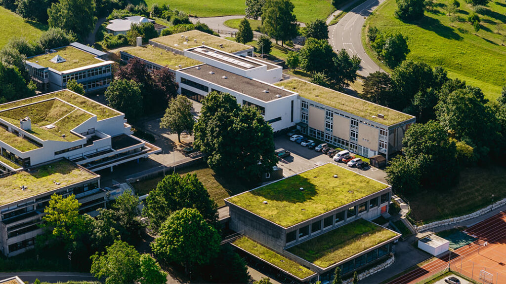 Luftaufnahme des Schulkomplexes der Messelbergschule mit mehreren Gebäuden, die flache Dächer mit begrünten Flächen haben, umgeben von Bäumen und einer Straße.