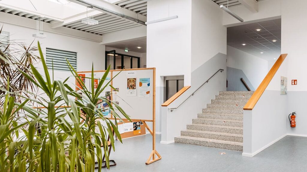 Innenraum eines Gebäudes mit Treppe, Pflanzen und einer Pinnwand mit Aushängen.