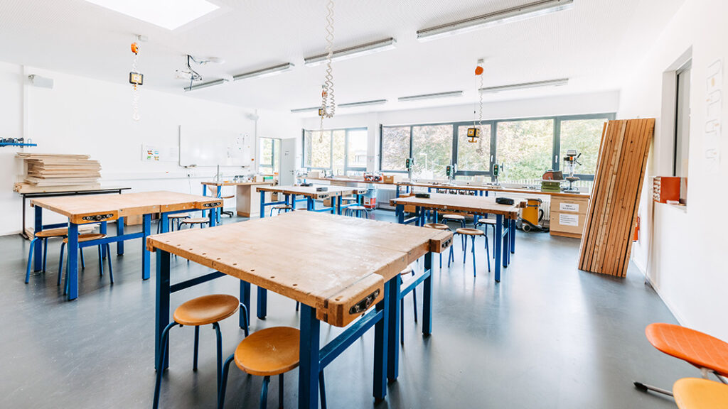 Großer heller Werkraum mit mehreren Arbeitstischen aus Holz und blauen Metallgestellen, Hockern und großen Fenstern an der Rückwand.