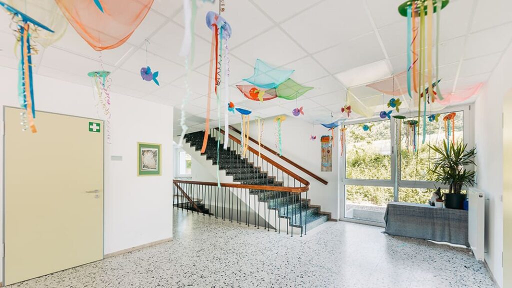 Heller Flur mit Treppe, großen Fenstern, mehreren bunten, netzartigen Dekorationen an der Decke und einer großen Pflanze neben einer Treppe.