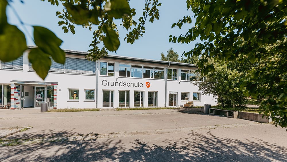 Zweistöckiges Schulgebäude mit der Aufschrift 'Grundschule' und einem Wappen neben dem Schriftzug, umgeben von Bäumen.