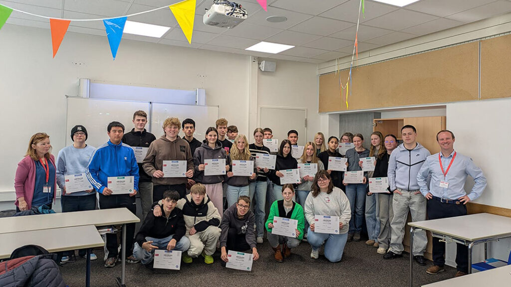 Gruppe von Personen in einem Klassenraum, die Zertifikate in den Händen halten, umgeben von bunten Wimpelketten an der Decke