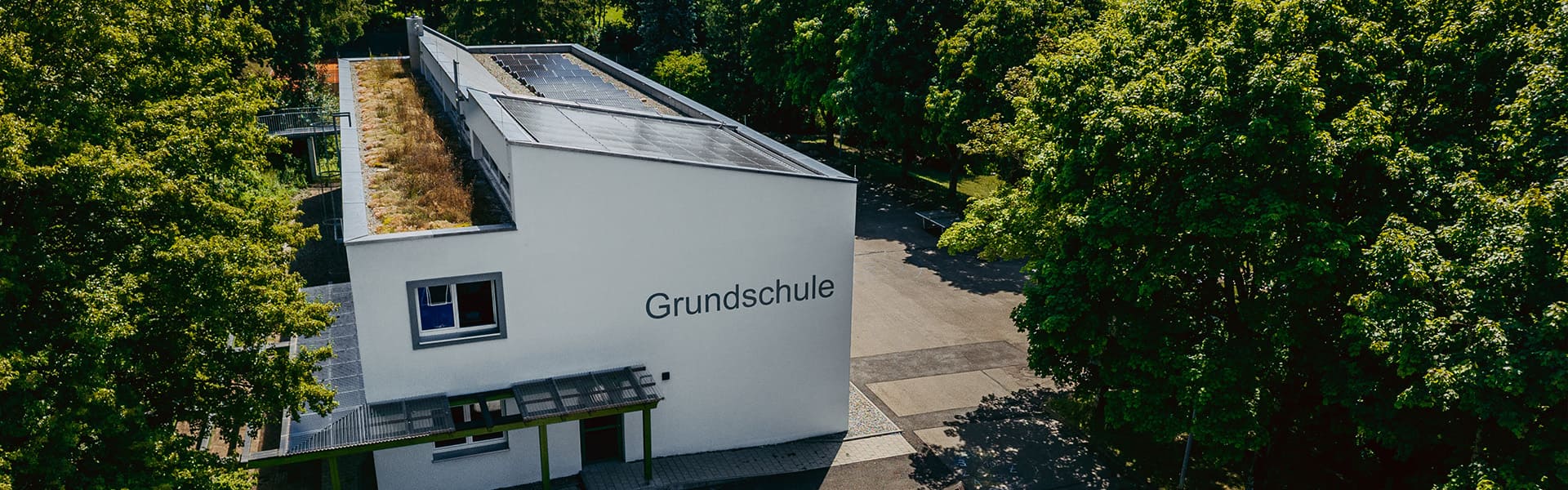 Eckansicht eines weißen Schulgebäudes mit der Aufschrift 'Grundschule' und einem Vordach, umgeben von Bäumen