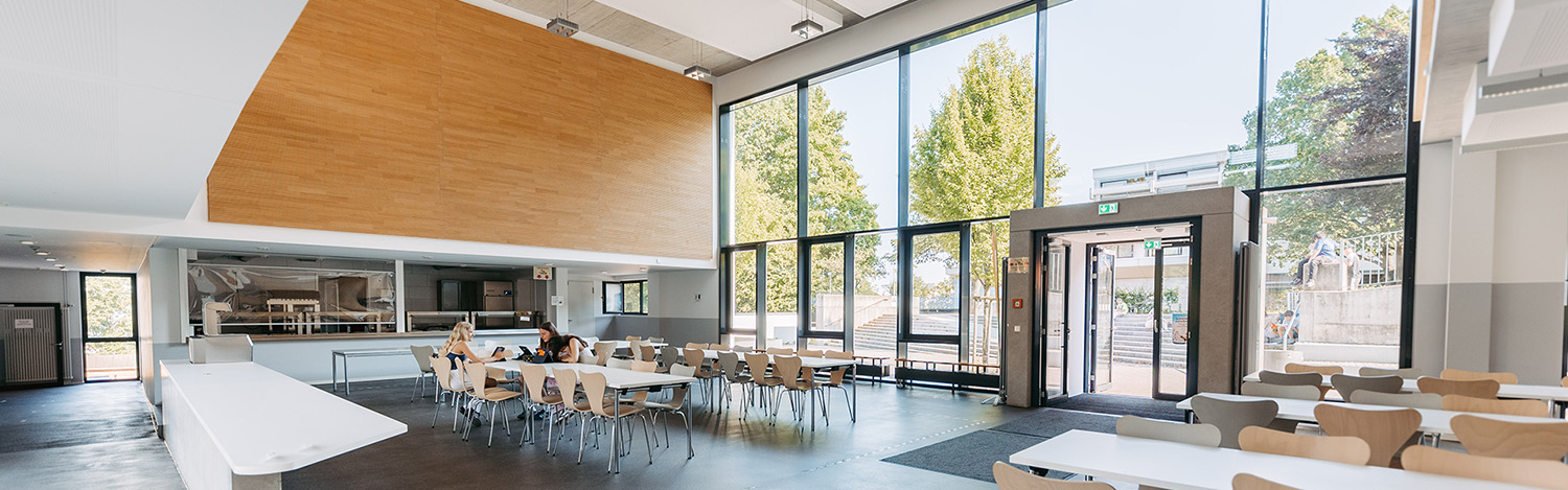 Großer heller Speisesaal mit mehreren Tischen und Stühlen vor großen Fenstern und Glastüren