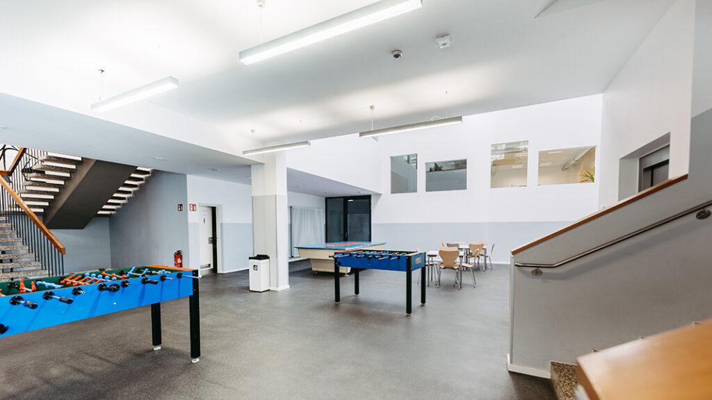 Großer heller Aufenthaltsraum mit blauem Tischkicker, Billardtisch, Sitzgruppe und Treppenaufgängen