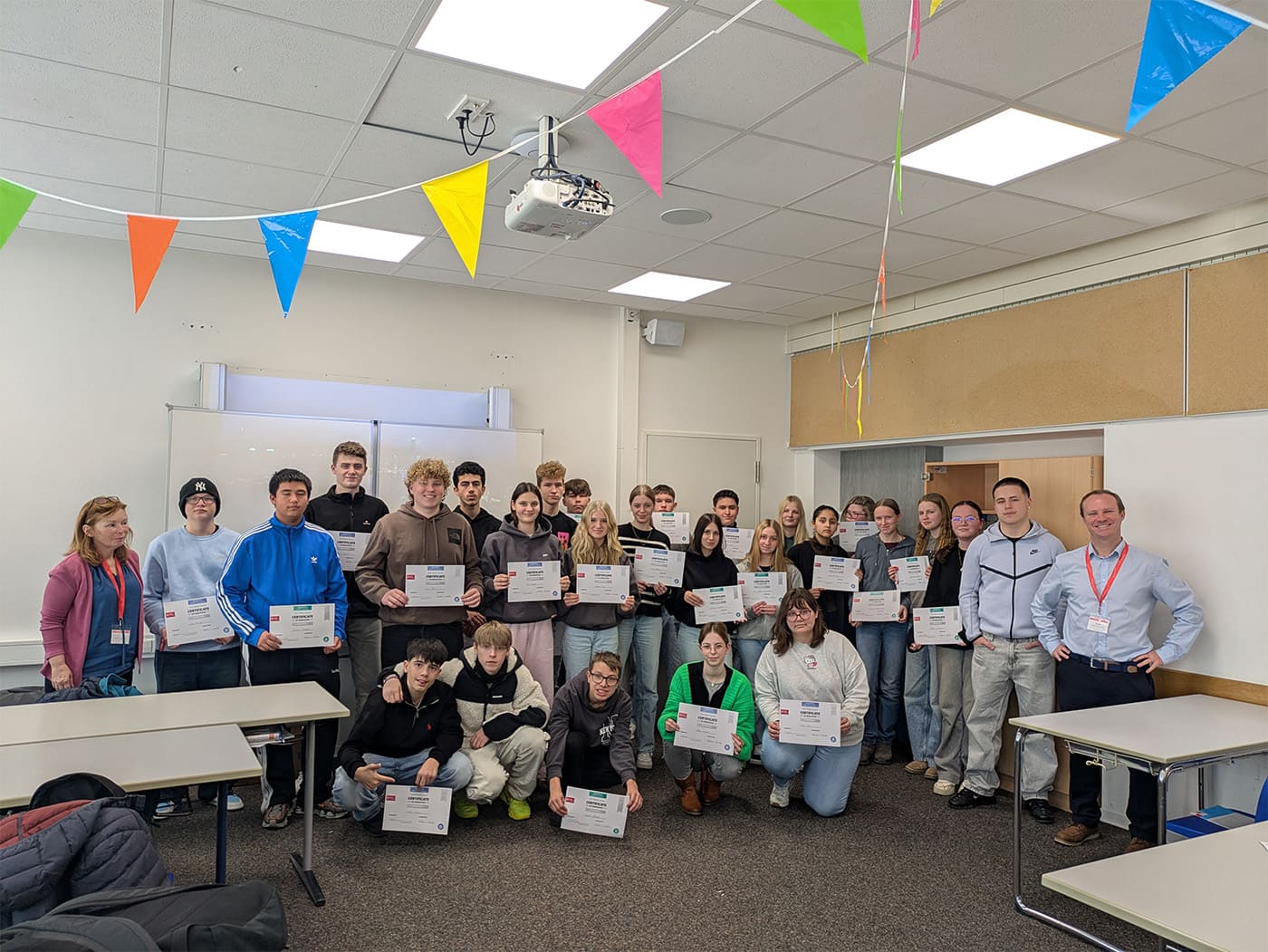 Gruppe von Personen in einem Klassenraum, die Zertifikate in den Händen halten, umgeben von bunten Wimpelketten an der Decke