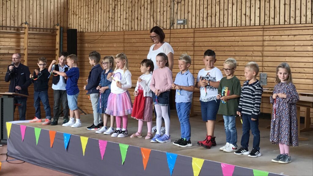 Kinder stehen in einer Reihe auf einer Bühne mit bunten Wimpeln am Bühnenrand, begleitet von einer erwachsenen Person im Hintergrund
