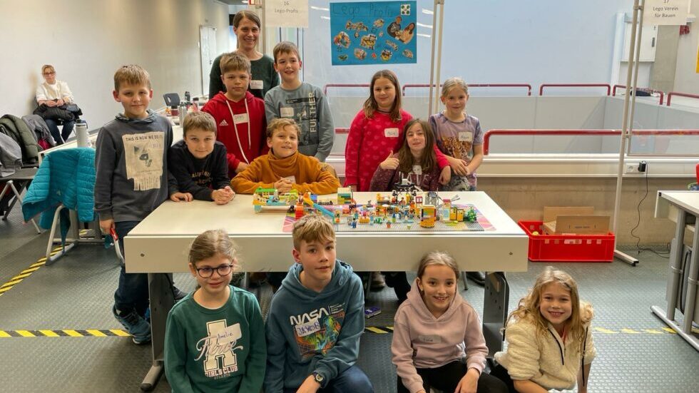 Gruppe von Kindern steht und sitzt um einen Tisch mit Lego-Modellen in einem hellen Raum mit Sitzbänken und Plakaten an der Wand