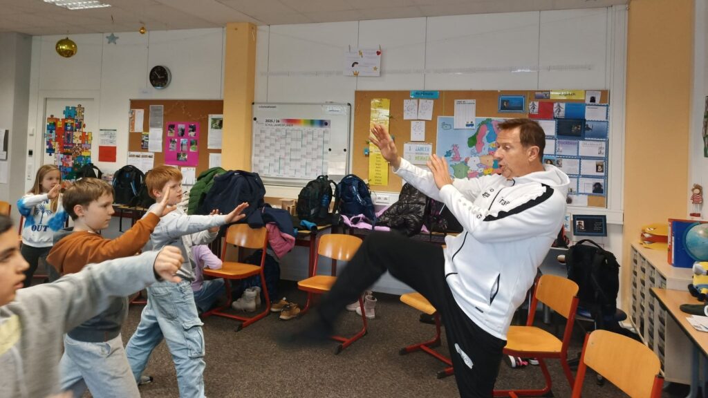 Mehrere Kinder und eine Person in Sportkleidung stehen in einem Klassenraum und strecken die Arme nach vorne