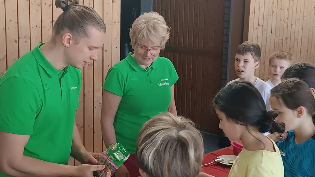 Gruppe von Kindern sitzt an einem Tisch in einem Raum mit Holzvertäfelung und zwei AOK-Mitarbeitenden in grünen Poloshirts stehen daneben