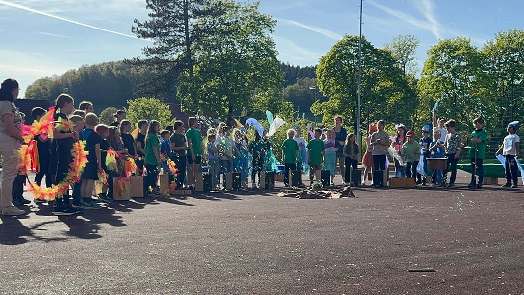 Gruppe von Kindern und Erwachsenen steht auf einem sonnigen Schulhof, einige Kinder tragen bunte Kostümteile und halten Musikinstrumente